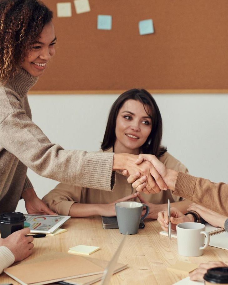 Five colleagues gathered around a conference table, smiling and talking while two in the center reach across to shake hands; coffee cups, notebooks, and a tablet with a colorful chart rest on the table, and a corkboard with sticky notes hangs in the background.