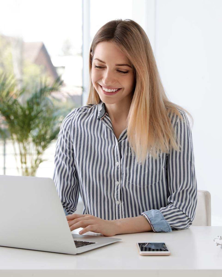 Smiling young woman in a striped shirt using a laptop at a bright desk with books and a smartphone nearby