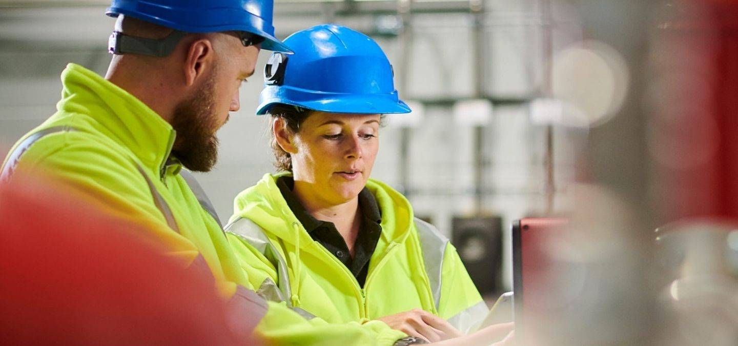 A man and woman in protective gear review data on a handheld device in an industrial facility.
