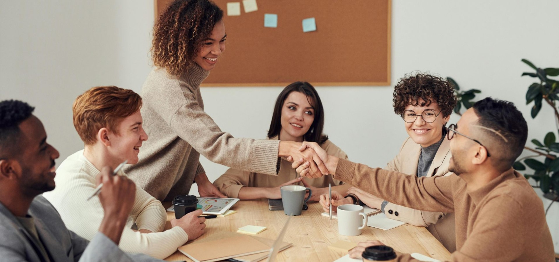 Five colleagues gathered around a conference table, smiling and talking while two in the center reach across to shake hands; coffee cups, notebooks, and a tablet with a colorful chart rest on the table, and a corkboard with sticky notes hangs in the background.