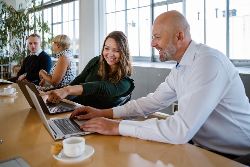 Smiling woman in a green sweater and a bald man in a white shirt work together at laptops in a bright office, with the man gesturing toward the keyboard.