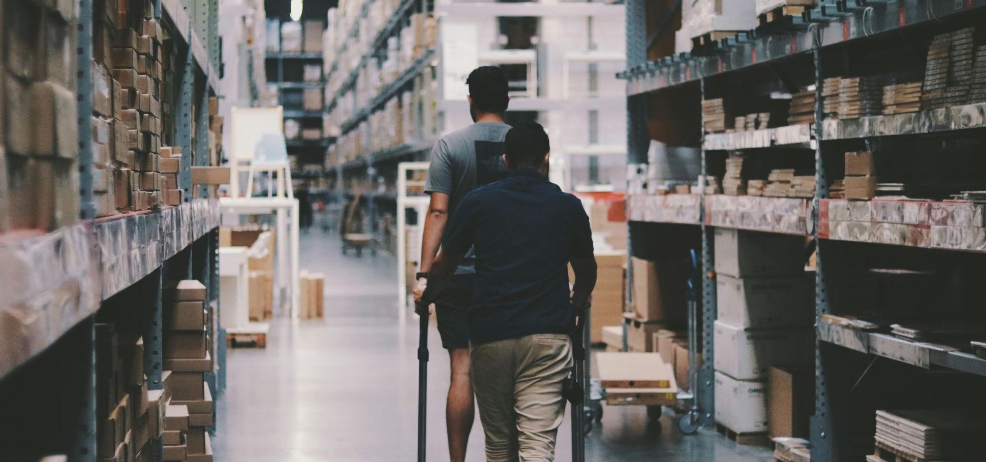 Two warehouse employees push a hand truck down a long aisle lined with tall shelving units stacked with cardboard boxes.