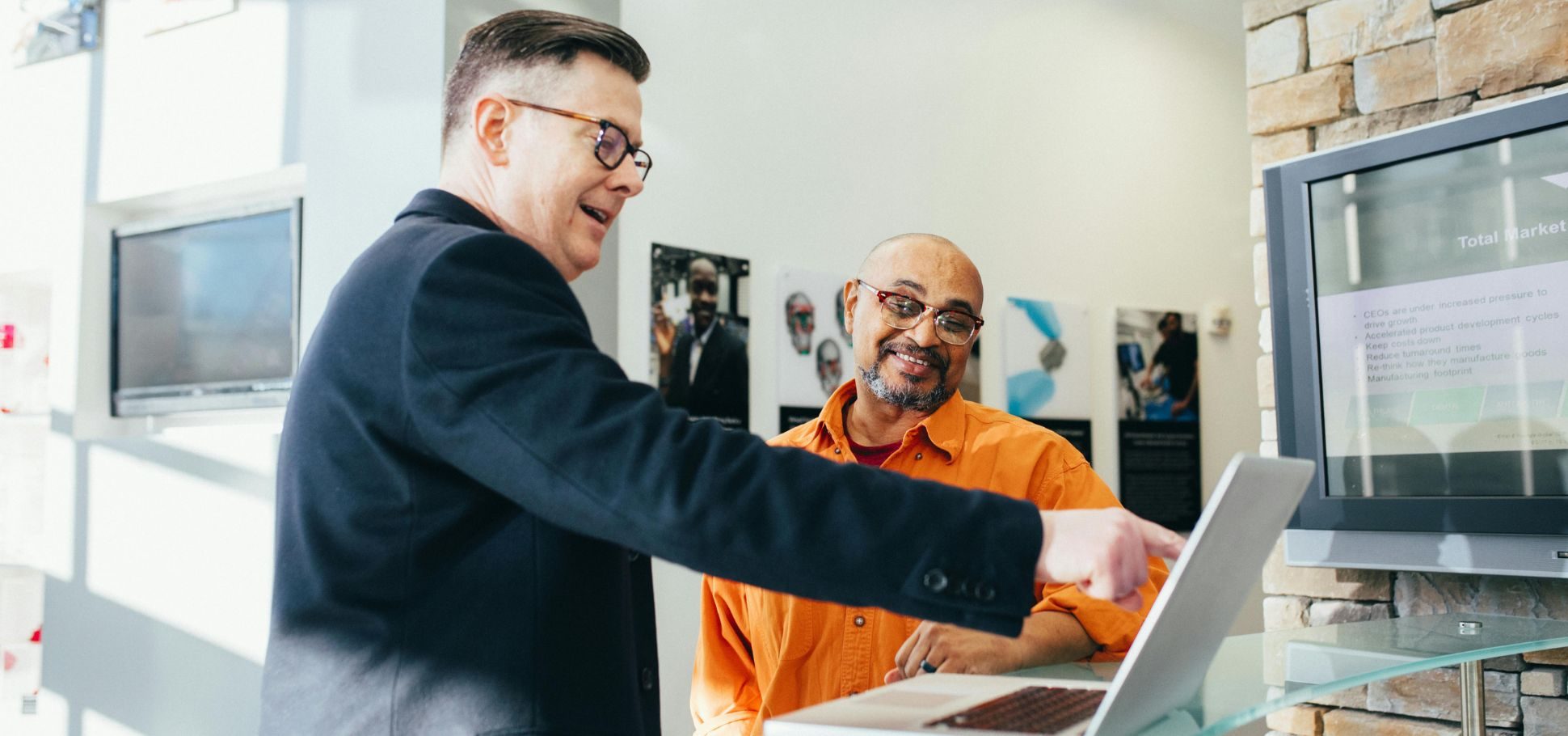 Two male business partners in a modern office, one in a dark suit pointing at a laptop screen while the other, wearing an orange shirt and glasses, smiles and listens. They are reviewing business data or a presentation slide on the computer and a large monitor in the background.