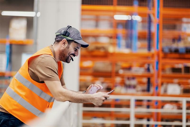 Warehouse worker in a safety vest smiling while looking at a mobile device.